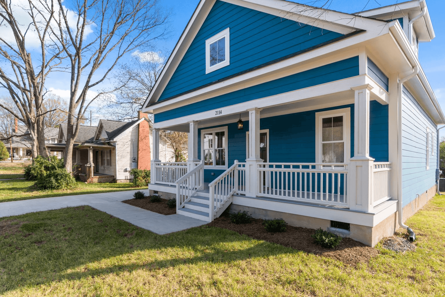 Photo of a blue two-story home with a white front porch, steps, sidewalk, lawn, tree, and neighboring house.